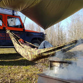 Person resting in a camo hammock under a Bushmen Thermo Tarp near a smoky campfire and Jeep.
